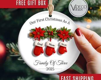 May include: A white ceramic Christmas ornament with a festive design. It features red mittens labeled "Dad," "Mom," and "Noah" hanging below a floral arrangement. The text reads "Our First Christmas As A Family Of Three 2025."