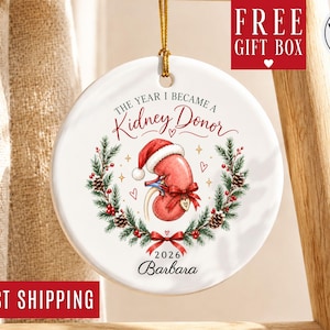 May include: A round white ceramic ornament featuring a red kidney illustration wearing a Santa hat, surrounded by a pine wreath with red berries and pinecones. Text reads: The Year I Became A Kidney Donor, 2026, and the name Barbara.