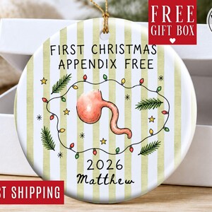 May include: A round ceramic Christmas ornament with a white and light green striped background. It features a central illustration of an appendix surrounded by a string of colorful holiday lights, pine sprigs, and stars. Text reads: FIRST CHRISTMAS APPENDIX FREE 2026 Matthew.