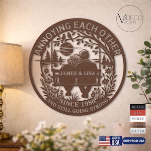 May include: A round, brown metal wall art piece with the text "ANNOYING EACH OTHER" and "AND STILL GOING STRONG SINCE 1986". It features a couple on a swing, with the names "JAMES & LISA" below. The design includes trees, flowers, and a sunset.