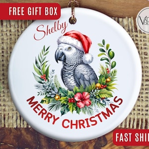 May include: White ceramic Christmas ornament featuring a gray parrot wearing a Santa hat, surrounded by greenery and red flowers. The ornament has the name "Shelby" and the words "Merry Christmas" in red.