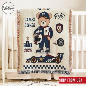 Peut inclure: Un plaid tissé de couleur crème représentant un ours en peluche vêtu d'une combinaison de course bleue et blanche tenant un casque. Le motif comprend une voiture de course, un drapeau à damier, un podium, le nom James Oliver et l'année 2026.
