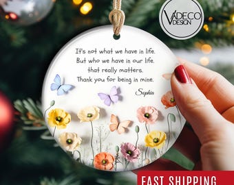 May include: A round, white ceramic ornament with a gold string for hanging. The ornament features a floral design with colorful poppies and butterflies. Text on the ornament reads: "It's not what we have in life...Thank you for being in mine. Sophia."