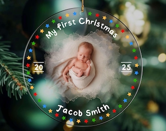 May include: A clear, round Christmas ornament with a photo of a newborn baby swaddled in a white blanket. The ornament is decorated with colorful stars and the text "My first Christmas" and "Jacob Smith". The numbers "20" and "25" are also displayed.