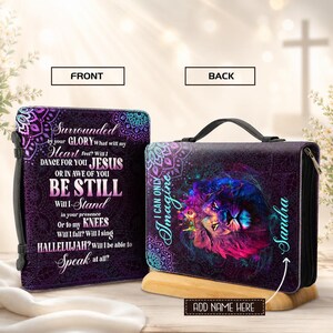 May include: A rectangular Bible cover with a black handle and zipper. The front features purple mandala patterns and text including "Surrounded by your GLORY" and "BE STILL". The back displays a colorful lion illustration and the name "Sandra".