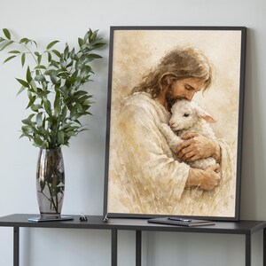 May include: A framed watercolor print depicts a figure with long brown hair and a beard, dressed in white robes, cradling a white lamb. The artwork has a soft, warm color palette and is set against a neutral background.