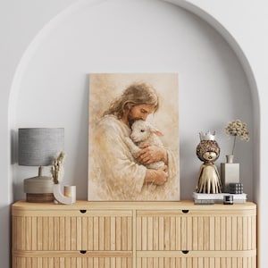 May include: A framed watercolor print depicts a figure with long hair embracing a lamb. The artwork is in soft, warm tones of beige and brown. The print is displayed on a wooden cabinet with a lamp and decorative items.