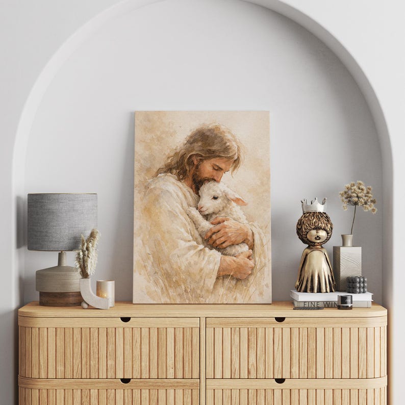 May include: A framed watercolor print depicts a figure with long hair embracing a lamb. The artwork is in soft, warm tones of beige and brown. The print is displayed on a wooden cabinet with a lamp and decorative items.