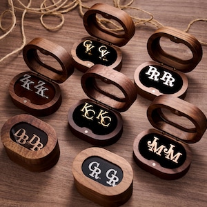 Peut inclure: Plusieurs boîtes à bijoux en bois avec des couvercles ovales sont ouvertes, révélant des boucles d'oreilles initiales. Les boucles d'oreilles sont dans des tons or et argent, sur fond de velours noir. Les boîtes sont disposées sur une surface en bois.