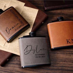May include: Three personalised leather flasks in shades of brown and grey, with black detailing. One flask is inscribed with the name "Damien" and the date "01.11.2025", another with "Dylan" and the date "04-08-2025", and the third with the initials "KM".