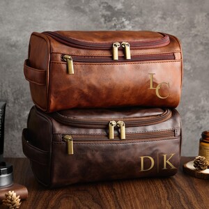 May include: Two brown leather travel bags with zipper closures and gold hardware. The top bag has the initials "LC" embossed on the front. The bottom bag has the initials "DK" embossed on the front.