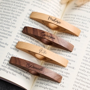 May include: Four wooden bookmarks with personalized names: Malarie, Georgia, Katie, and Danielle. The bookmarks are in various wood tones, including light and dark brown, and are designed to hold a book open. The names are written in elegant script.