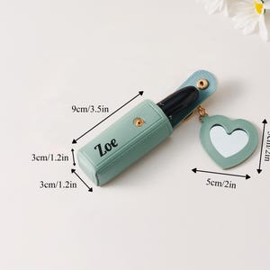 May include: A light green lipstick case with the name Zoe printed in black, featuring a gold snap closure and a heart-shaped mirror charm. The case measures 3.5 inches long by 1.2 inches wide and deep, with a 2-inch heart mirror charm attached.