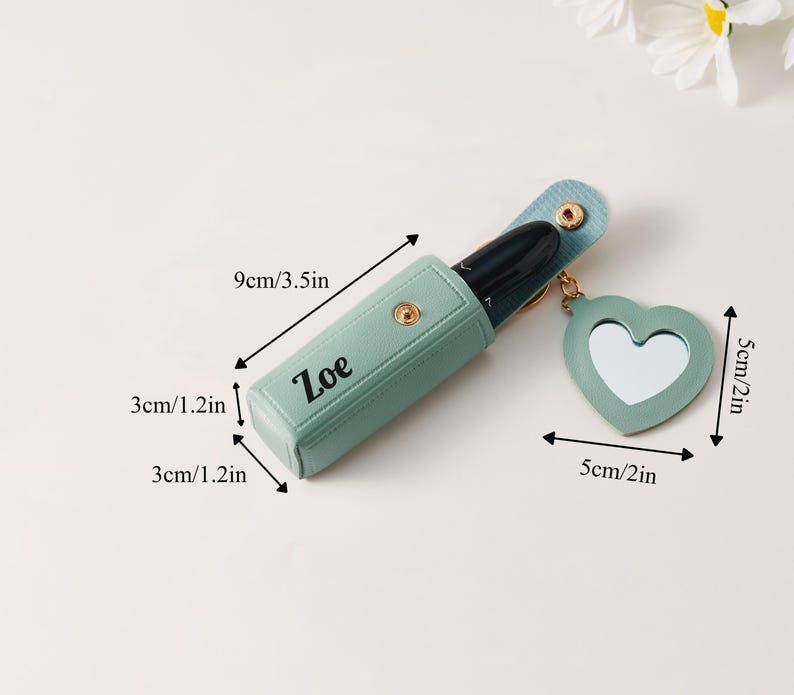 May include: A light green lipstick case with the name Zoe printed in black, featuring a gold snap closure and a heart-shaped mirror charm. The case measures 3.5 inches long by 1.2 inches wide and deep, with a 2-inch heart mirror charm attached.