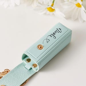 May include: A light mint green rectangular lipstick holder with a gold snap closure and the name Nicole printed in black script. The holder features a matching strap and a gold metal keyring attachment, showcasing a textured finish throughout.