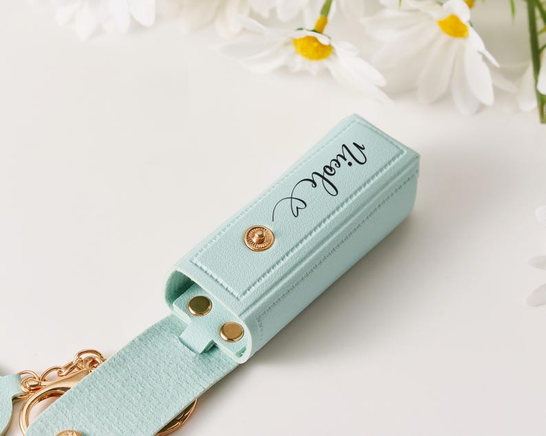 May include: A light mint green rectangular lipstick holder with a gold snap closure and the name Nicole printed in black script. The holder features a matching strap and a gold metal keyring attachment, showcasing a textured finish throughout.