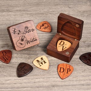 Può includere: Una scatola di legno con un design di chitarra sul coperchio e un plettro all'interno. La scatola è circondata da sei plettri per chitarra con diversi design e nomi.