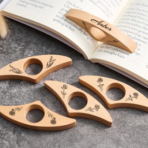 May include: A collection of wooden thumb page holders with floral designs and a personalized one with the name "Amber". The page holders are light brown and are resting on a book and a gray surface.