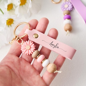 May include: Pink leather keychain with the name "Layla" in script. The keychain features a gold ring, a pink flower charm, and a beaded accent with wooden, pink, and white beads. A second keychain with a purple flower and beads is also visible.