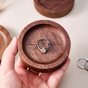 Könnte beinhalten: Eine runde Ringbox aus dunklem Holz mit einem Diamant-Verlobungsring im Inneren. Die Box hat einen geschnitzten Innenraum mit einem personalisierten Design. Zwei silberne Ringe liegen neben der Box. Die Box wird von einer Hand gehalten.
