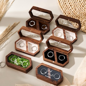 May include: A collection of wooden ring boxes, some open to show rings. The boxes are hexagonal and rectangular, with clear lids and personalized text. Some have moss or black velvet interiors. The rings are silver.
