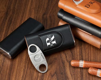 May include: A collection of leather cigar cases in black, brown, and tan, with a silver cigar cutter. The cases are personalized with initials and names. A cigar is visible in the foreground.