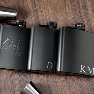 May include: Three black metal hip flasks with engraved lettering. The flasks are on a wooden surface. The first flask says "Dylan Groomsm 04-08-20". The second flask says "D". The third flask says "KM".