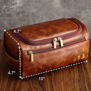 May include: A brown leather travel toiletry bag with two zippers and a gold zipper pull. The bag measures 10 inches long, 4.7 inches wide, and 4.7 inches high. The bag has a monogrammed initial "M" on the front.