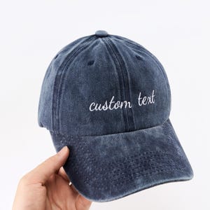Op de afbeelding: Marineblauwe baseballcap met de woorden "custom text" in witte letters geborduurd. De cap heeft een gebogen klep en een stoffen bandsluiting. Gemaakt van zacht, gewassen katoen.