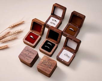 May include: Several open wooden ring boxes displaying silver rings. Some boxes have red or brown velvet interiors. Two boxes are engraved with names and dates. The boxes are arranged on a light-coloured surface.