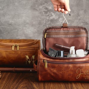 May include: Two brown leather travel bags with zipper closures. The bag on the left has the name "Mark" embossed on the front. The bag on the right is open and has a mesh pocket inside. The bag on the right has the name "Justin" and the date "08-05-2023" embossed on the front.