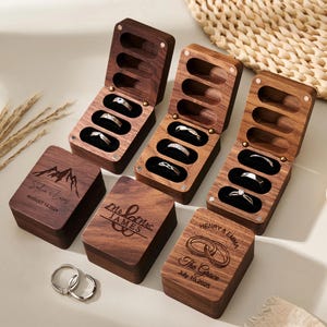 Op de afbeelding: Houten ringendoosjes met zilveren ringen erin, sommige open en sommige gesloten. De dozen zijn gegraveerd met namen en data. Twee zilveren ringen staan op de voorgrond. De dozen zijn gemaakt van donker hout.