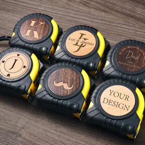 Può includere: Sei metri a nastro gialli e neri con inserti in legno personalizzati. Gli inserti presentano diversi design, tra cui iniziali, un baffo e la parola "Papà".