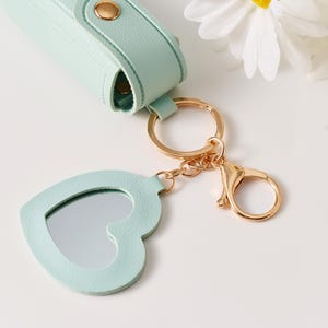 May include: A light mint green lipstick case with a gold snap closure attached to a gold keyring and a heart-shaped mirror charm. The mirror is framed in matching mint green material and connects to the keyring via a gold chain and lobster clasp.