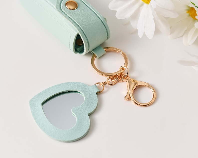 May include: A light mint green lipstick case with a gold snap closure attached to a gold keyring and a heart-shaped mirror charm. The mirror is framed in matching mint green material and connects to the keyring via a gold chain and lobster clasp.