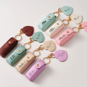 May include: A collection of rectangular lipstick cases with snap closures, each featuring a gold-tone keyring and a heart-shaped charm. The cases are in various pastel and solid colors, including mint, pink, purple, cream, and maroon, with names printed.