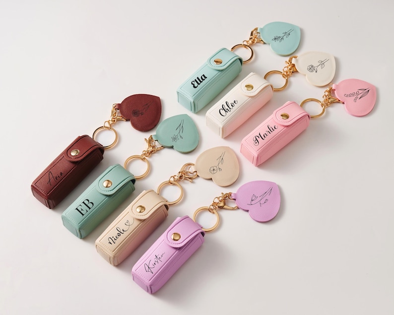 May include: A collection of rectangular lipstick cases with snap closures, each featuring a gold-tone keyring and a heart-shaped charm. The cases are in various pastel and solid colors, including mint, pink, purple, cream, and maroon, with names printed.