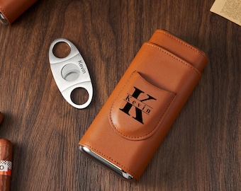 May include: A brown leather cigar case with a silver trim and a pocket featuring the name "Kevin" in black. A silver cigar cutter with the name "Kevin" is next to the case. The items are on a wooden surface.