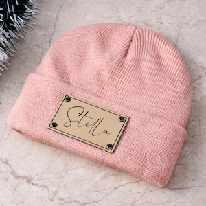 Op de afbeelding: Een roze gebreide beanie met een bruin leren patch waarop "Stella" in cursief staat.