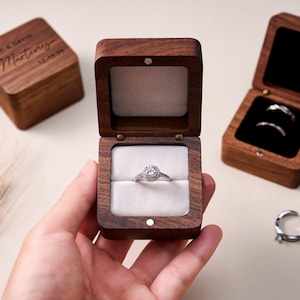 Op de afbeelding: Een zilveren verlovingsring met een halo-zetting wordt getoond in een open, vierkante, houten ringdoos. Twee andere houten ringdozen zijn ook zichtbaar, één gesloten en één open met ringen erin. De gesloten doos heeft de inscriptie "JANE & DAVID Martinez 12.19.24".