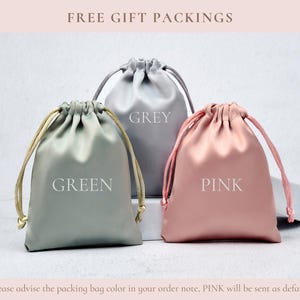 May include: Three satin drawstring pouches in green, grey, and pink. Each pouch features a matching drawstring closure and the color name printed in white text on the front. The green pouch has gold cords, while the grey and pink pouches have matching cords.