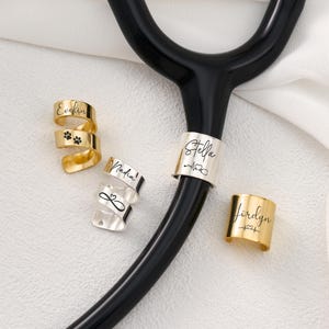 Op de afbeelding: Een verzameling gepersonaliseerde stethoscoopbedels in goud en zilver. De bedels zijn gegraveerd met namen, pootafdrukken en medische symbolen. De stethoscoop is zwart en de achtergrond is wit.