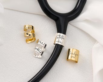 May include: A collection of personalized stethoscope charms in gold and silver. The charms are engraved with names, paw prints, and medical symbols. The stethoscope is black, and the background is white.