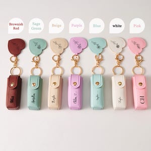 May include: Seven rectangular lipstick cases with gold-tone snap closures and matching heart-shaped keychains, each featuring a unique floral line drawing and a personalized name or initials in black text, available in brownish red, sage green, beige, purple, blue, white, and pink.