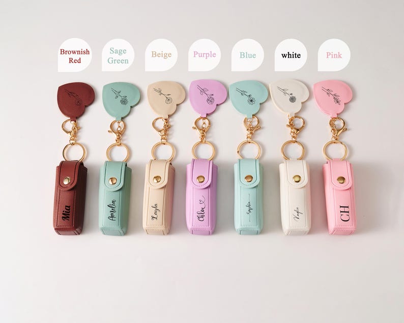 May include: Seven rectangular lipstick cases with gold-tone snap closures and matching heart-shaped keychains, each featuring a unique floral line drawing and a personalized name or initials in black text, available in brownish red, sage green, beige, purple, blue, white, and pink.