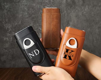 May include: Three leather cigar cases in black, brown, and tan. Each case has a silver cigar cutter and a pocket with personalized initials or a name. The cases are cylindrical and appear to be made of high-quality leather.