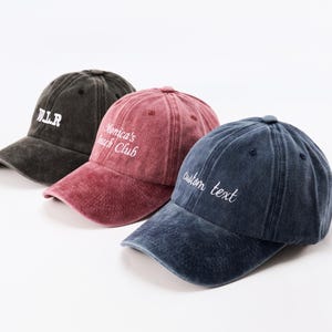May include: Three baseball caps in a row, each with a different colour and embroidered text. The caps are in dark grey, maroon, and navy blue. The text on the caps reads "W.L.R", "Monica's Beach Club", and "custom text".