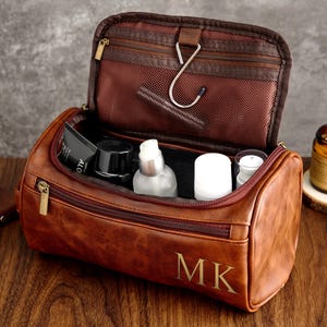 May include: Brown leather travel toiletry bag with a zipper closure and a hook inside. The bag is open and filled with toiletries, including a bottle of lotion, a tube of toothpaste, and a small bottle of shampoo. The bag has the initials "MK" embossed on the front.