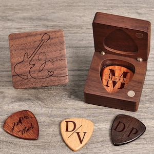 May include: A wooden box with a guitar design on the lid and the name "Jason" engraved. The box contains a guitar pick with the name "Mark" engraved. There are three additional guitar picks with different designs and engravings.