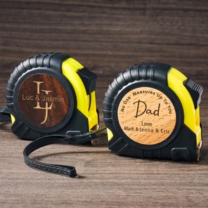 May include: Two yellow and black tape measures with wooden engraved faces. One reads "Luc & Jasmin" with a monogrammed "LJ". The other reads "No One Measures Up To You Dad Love Mark & Jenna & Eric".